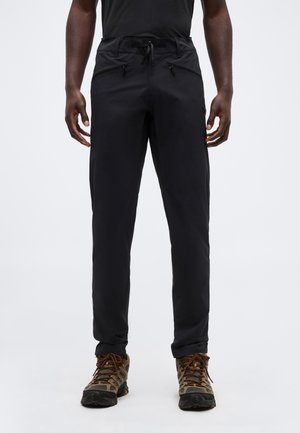 Άντρας που φοράει μαύρο παντελόνι με φερμουάρ και καφέ παπούτσια πεζοπορίας, στέκεται μπροστά σε λευκό φόντο, με θέα από τη μέση μέχρι τα πόδια.