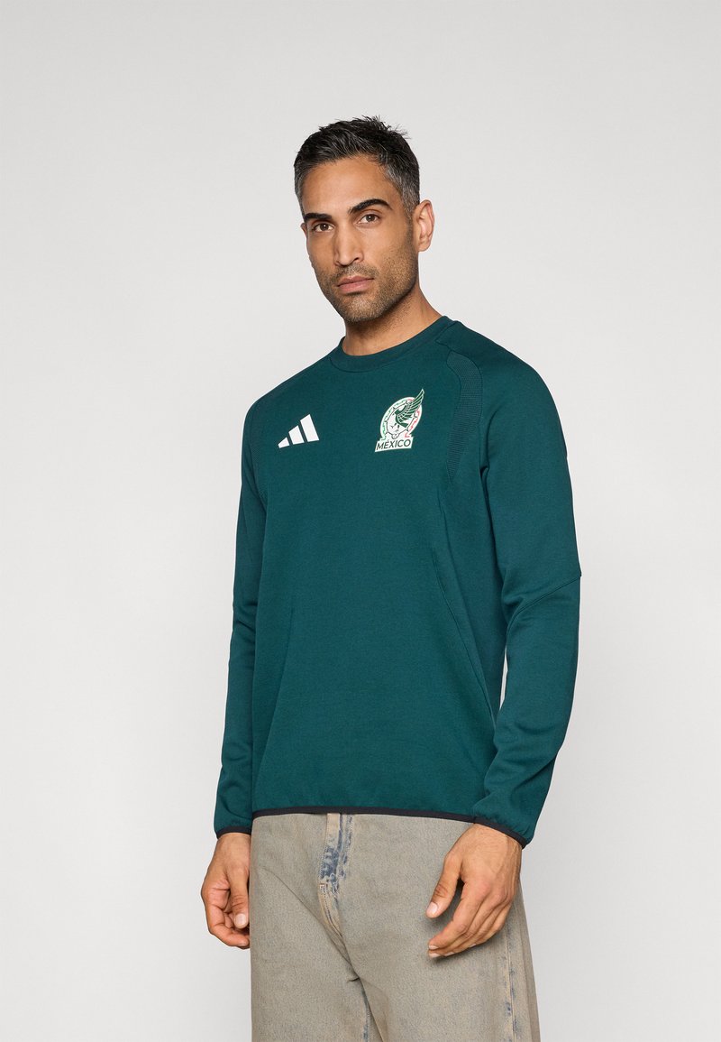 Πράσινο αθλητικό μπλουζάκι με μακριά μανίκια και στρογγυλή λαιμόκοψη, με λευκό λογότυπο και τρεις λευκές ρίγες στο αριστερό στήθος.