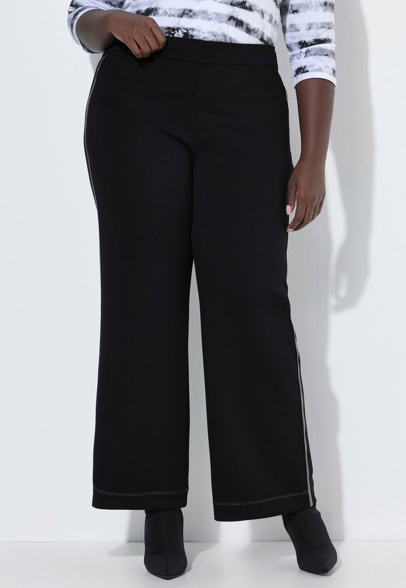 Kobieta ubrana w czarne spodnie o szerokich nogawkach z bocznym paskiem, stojąca na tle białej ściany, w połączeniu z czarnymi, szpiczastymi botkami na kostkę.