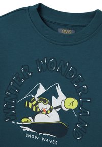 Cyan sweatshirt met een cartoon sneeuwman die snowboardt met een gestreepte sjaal. Tekst leest "WINTER WONDERLAND" en "SNOW WAVES."