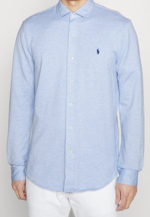 Chemise bleu clair à manches longues avec col et boutons, logo de joueur de polo marine sur la poitrine gauche, portée par une personne portant un pantalon blanc.