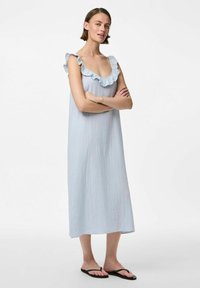 Gaiši zila bez piedurknēm kleita ar volāniem ap kaklu un teksturētu audumu, kas sasniedz puslīdz ceļgaliem. Valkāta ar melnām flip-flop sandalēm.