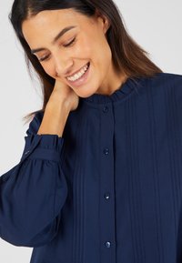 Blusa blu navy con colletto arricciato, dettagli a righe verticali e chiusura con bottoni. Presenta maniche a sbuffo e tessuto testurizzato.
