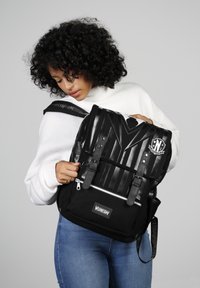 Sac à dos noir avec un devant texturé, des bretelles ajustables, deux boucles, une poche zippée et un patch logo, adapté à un usage quotidien.