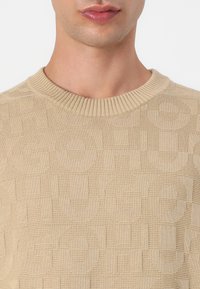 Beige kudumüts sisaldab tekstureeritud mustrit, mille sees on brändinimi "HUGO" erinevates suurustes. Sisaldab klassikalist soonikserva kaelaosa.