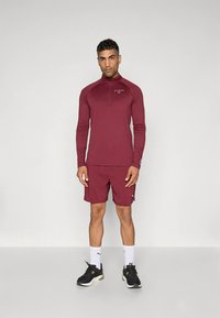 Conjunto de roupa desportiva cor bordô, composto por uma camisola de manga comprida com fecho de correr e calções combinando. O tecido parece macio, com uma marca subtil e acentos contrastantes.