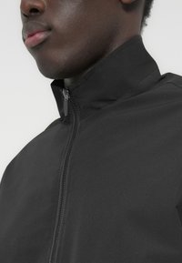 Μαύρο μπουφάν με φερμουάρ και ψηλό γιακά, με λεία υφή, διαθέτει μπροστινό φερμουάρ και μινιμαλιστικό σχέδιο.