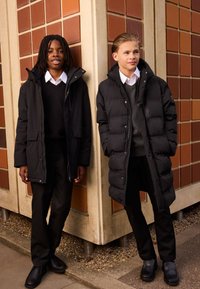 Deux jeunes portent des vestes matelassées noires avec capuches, associées à des pantalons noirs et des chaussures vernies, devant un mur géométrique texturé.