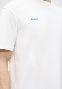 Λευκό κοντομάνικο μπλουζάκι με μικρό μπλε λογότυπο «ARKK» στο αριστερό στήθος, φορεμένο από άτομο με ανοιχτόχρωμο δέρμα σε απλό φόντο.