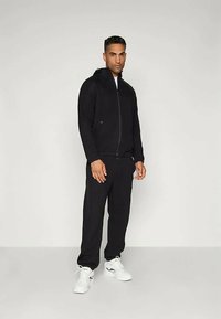 Μαύρο αθλητικό σύνολο με ζακέτα με φερμουάρ και ταιριαστά παντελόνια, με λείο ύφασμα και εφαρμοστή σχεδίαση. Λευκά αθλητικά παπούτσια ολοκληρώνουν την εμφάνιση.