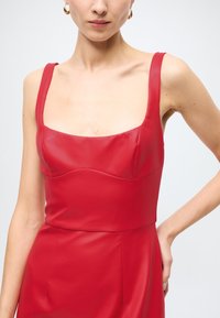 Vestido vermelho sem mangas com decote quadrado, feito de tecido liso e brilhante. Apresenta um corpete ajustado e um ligeiro franzido ao longo das costuras.