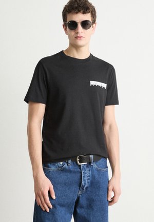 T-shirt di cotone nero con maniche corte, caratterizzata da un logo bianco di Calvin Klein sul lato sinistro del petto. Indossato con jeans in denim blu e una cintura nera.