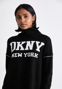 Svart stickad tröja med hög ribbad krage. Har vit text "DKNY NEW YORK" och kontrasterande horisontell sömdetalj.