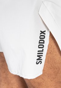 Shorts blancos de tejido ligero que presentan un logo negro "SMILODOX" en el lateral. El diseño incluye una abertura y una textura suave.