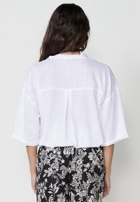Blusa bianca in lino a taglio ampio con maniche corte e piega sul retro, abbinata a pantaloni neri con motivo floreale.