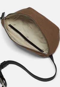 Brun lærred crossover taske med sort læderrem, delvist åben sølvlynlås, der viser beige indvendig foer med mærkets logo og indvendig lynlås lomme.