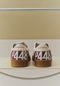 Paire de baskets blanches et marron avec un grand texte blanc "P448" et "You Can Surf Later" écrit sur les languettes de talon, exposées sur une étagère claire.