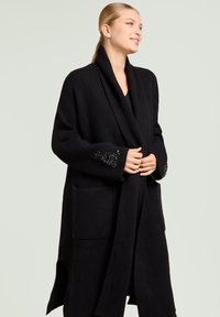 Zwarte, lange wollen jas met een sjaalkraag, grote voorzakken en versierde manchetdetails. Glad oppervlak, open voorkant ontwerp.