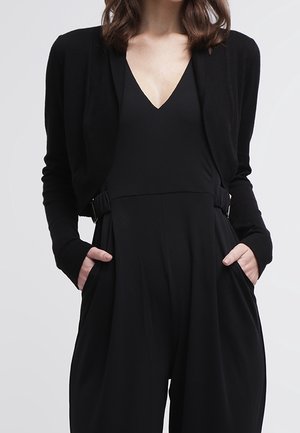 Schwarze Strickjacke mit langen Ärmeln, die über einem schwarzen Jumpsuit getragen wird. Die Strickjacke hat einen offenen Vorderteil und eine weiche Textur; der Jumpsuit hat weit geschnittene Beine und Taschen.