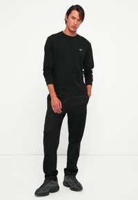Jonge man in een zwart overhemd met lange mouwen en zwarte broek met zwarte sneakers tegen een eenvoudige witte achtergrond.