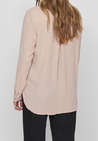 Femme aux longs cheveux bruns portant un chemisier ample à manches longues rose clair et un pantalon noir, vue de dos sur un fond uni.