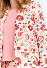 Blazer à motif floral en crème avec des fleurs rouges, roses et bleues ; doté d'un col cranté et d'une fermeture à un bouton. En dessous, un top rose.