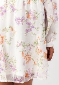 Robe florale blanche en tissu léger, avec des motifs de fleurs orange, violets et verts, des manches bouffantes et un ourlet plissé.