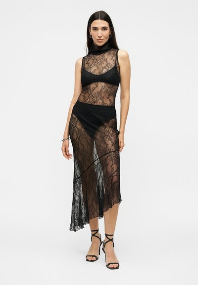 Femme portant une robe noire en dentelle sans manches avec un col roulé et des sous-vêtements, associée à des talons hauts noirs à brides, debout devant un fond blanc.