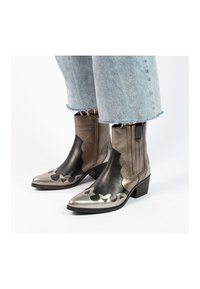 Metallic bronze cowboy boots with pointed toes, decorative cut-outs, and ribbed elastic sides. Chunky black heel and smooth texture.
