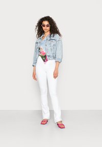 Casaco de ganga azul claro, top gráfico floral colorido, jeans brancos ajustados e sandálias planas cor-de-rosa, com óculos de sol e cabelo encaracolado.