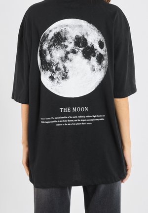 Persoon draagt een zwart t-shirt met een grote witte gedetailleerde maanafbeelding en beschrijvende tekst over de maan op de achterkant.