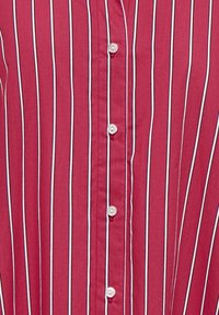 Chemise rouge à rayures verticales blanches et noires, avec une rangée centrale de cinq boutons blancs.
