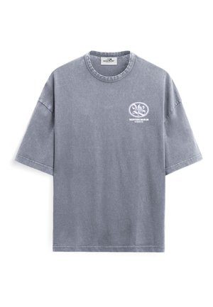Osoba z krótkimi włosami, ubrana w wyblakły szary T-shirt z białymi gwiazdami i napisem "Vertere Berlin Services", stojąca na jednolitym tle.