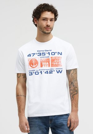 Mann mit lockigem Haar, der ein weißes T-Shirt mit nautischen Koordinaten, orangefarbenem Anker und Segelboot-Grafiken sowie blauem Text trägt, steht vor schlichtem Hintergrund.