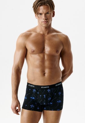 Młody, muskularny mężczyzna bez koszulki, ubrany w czarne bokserki z niebieskim kwiatowym nadrukiem i paskiem z napisem "ATLANTIC", stojący na białym tle.