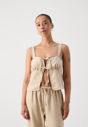Beige cropped top with a tie front, adjustable straps, and a relaxed fit. Material appears soft with a smooth texture.