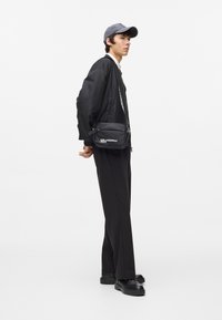 Jachetă bomber neagră, pantaloni negri și pantofi negri. Capt de culoare gri și o geantă transversală mică, neagră, cu text alb pe ea. Design minimalist.