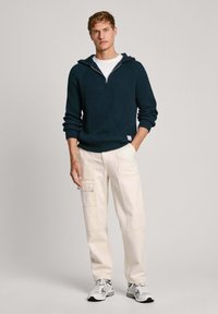 Tamsiai mėlynas lankstus džemperis su užtrauktuku, dėvimas ant baltos marškinėlių. Derinamas su kremiginėmis kelnėmis su kišenėmis ir laisvu siluetu. Matomos sportinės bateliai.