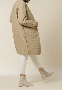 Beige quilted coat with long sleeves, paired with white cropped jeans and light grey, textured ankle boots with block heels.