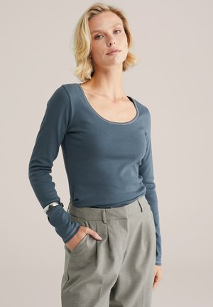 Frau mit kurzem blonden Haar, die ein enges blaues Langarmoberteil und hoch geschnittene graue plissierte Hose trägt, mit einer Hand in der Tasche.