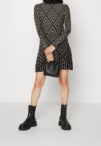 Femme portant une robe longue à manches longues noire et beige à motifs, tenant un petit sac à main noir, accompagnée de bottines noires à semelle épaisse.