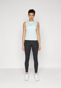 Svetlo zelena brezrokavnik s belim logotipom Puma, v paru z črnimi legicami s stranskimi poudarki. Model nosi bele športne čevlje.
