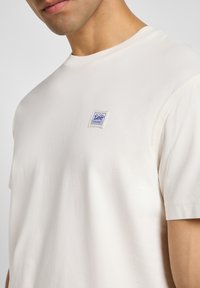 T-shirt crema a maniche corte in morbido cotone con colletto rotondo, caratterizzata da una piccola etichetta con logo Lee blu e bianco sul petto.