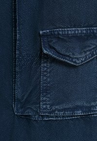 Tummansininen farkkukangas, jossa on teksturoitu pinta, varustettuna yhdellä läpällisellä taskulla ja kontrastivärisellä ompelukohdalla reunoissa.