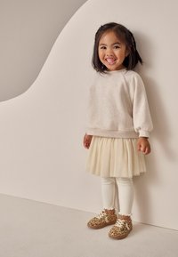 Ljusbeige tröja, krämfärgad plisserad kjol, vita rippade strumpbyxor och skor med leopardtryck. Barnet står mot en minimalistisk bakgrund.