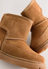 Ghete de gleznă din piele întoarsă maro, cu vârf rotund, cusături vizibile și talpă din cauciuc texturată. Marginea superioară este căptușită cu un material textil moale.