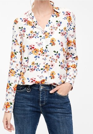Vrouw draagt een wit blouse met lange mouwen en een kleurrijke bloemenprint, ingestopt in een blauwe spijkerbroek, met één hand in de zak.
