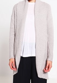 Ljusgrå stickad cardigan med öppen framsida, texturerat tyg, lång ärm och avslappnad passform, lager på lager över en vit skjorta.