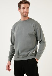 Sudadera gris hecha de un tejido suave, con un cuello redondo, mangas largas, puños acanalados y un pequeño parche con logo en el dobladillo.