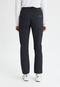 Person i svarta raka byxor och vita Nike Air-sneakers, stående vänd bort mot en enkel vit bakgrund.